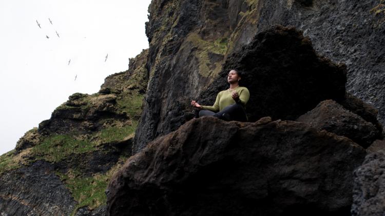Person in Zen pose outside a cave with sky behind them.